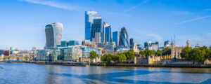 London skyline with modern skyscrapers and historic landmarks along River Thames. Iconic buildings like the Gherkin, Walkie-Talkie, and Tower of London visible under blue sky.