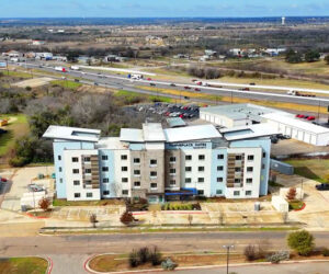 Aerial view of a TownePlace Suites hotel near a major highway. The light blue building stands out against the surrounding rural landscape, with a busy road and open fields visible.