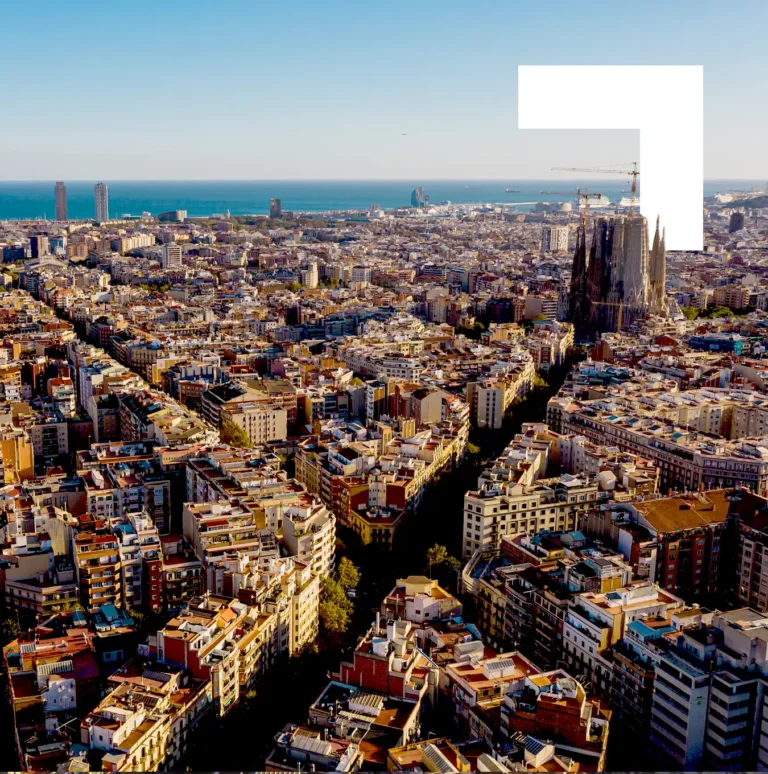 Aerial view of Barcelona showing the dense urban landscape with Sagrada Familia visible, Mediterranean coastline in the background, and the city's distinctive grid pattern of streets under clear blue skies.